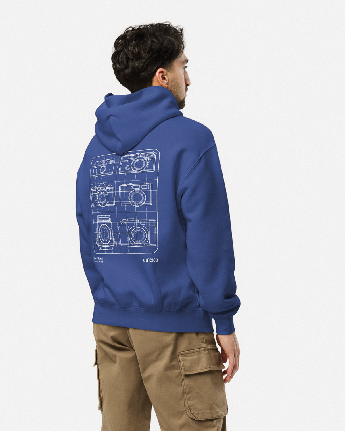 Photo of a man wearing a blue cineica "Blueprint Hoodie". The white design showcases six iconic analog film cameras and the text "Analog 'Blueprint", "35mm or 120 Film".