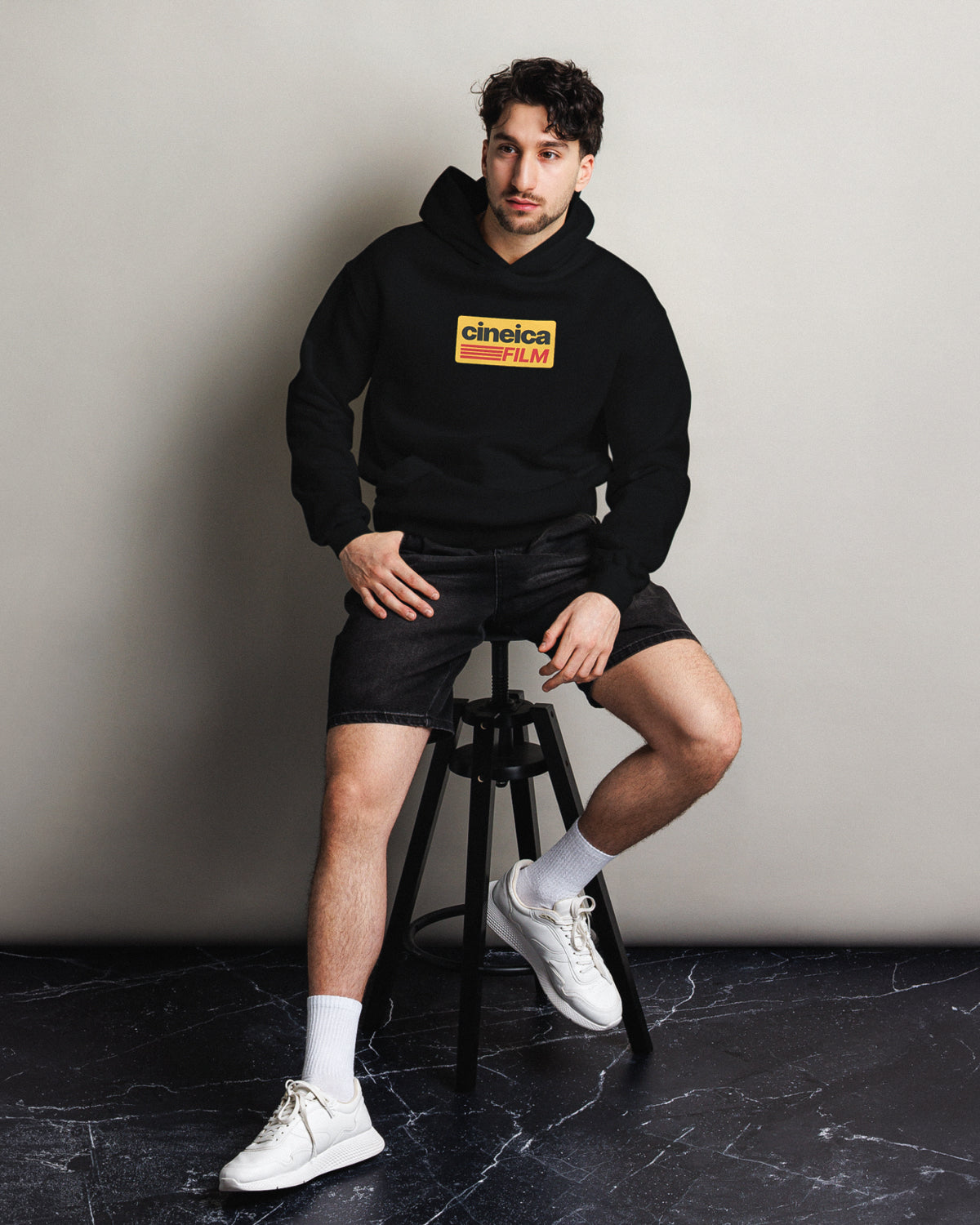 Photo of a man sitting on a stool wearing a black cineica hoodie. The hoodie design features vintage colors and text "cineica film" in a Kodak film style.