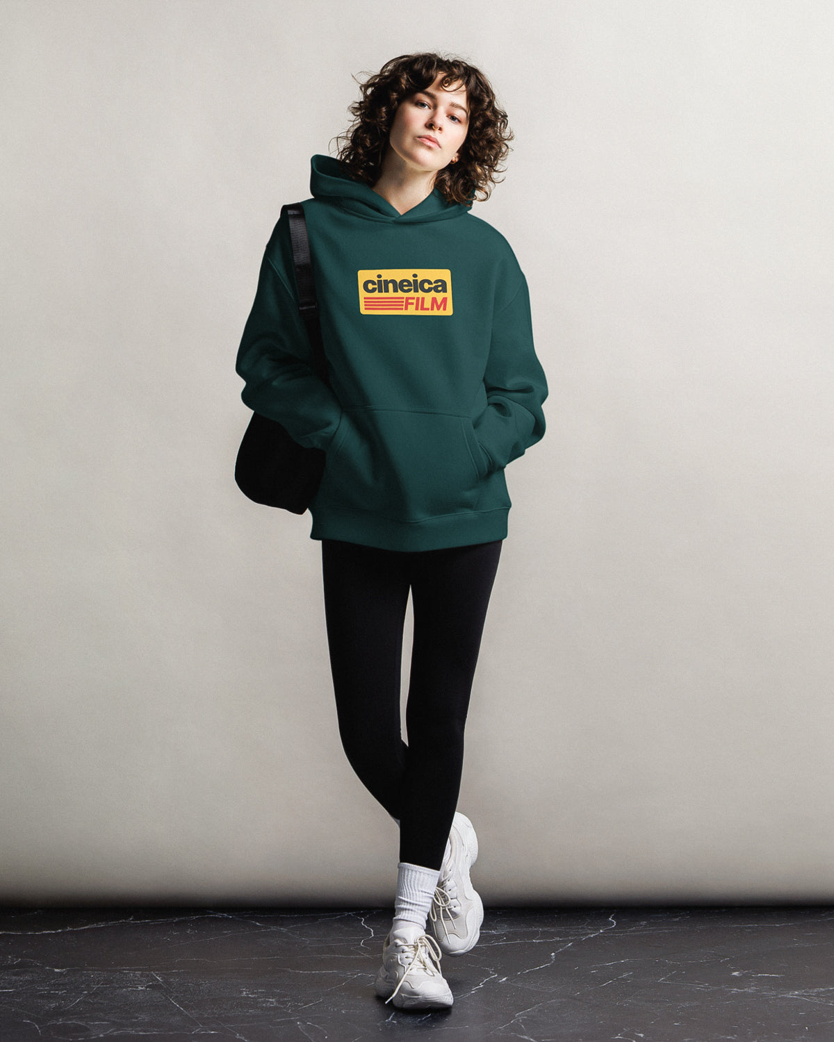 Photo of a woman wearing a green cineica hoodie. The hoodie design features vintage colors and text "cineica film" in a Kodak film style.